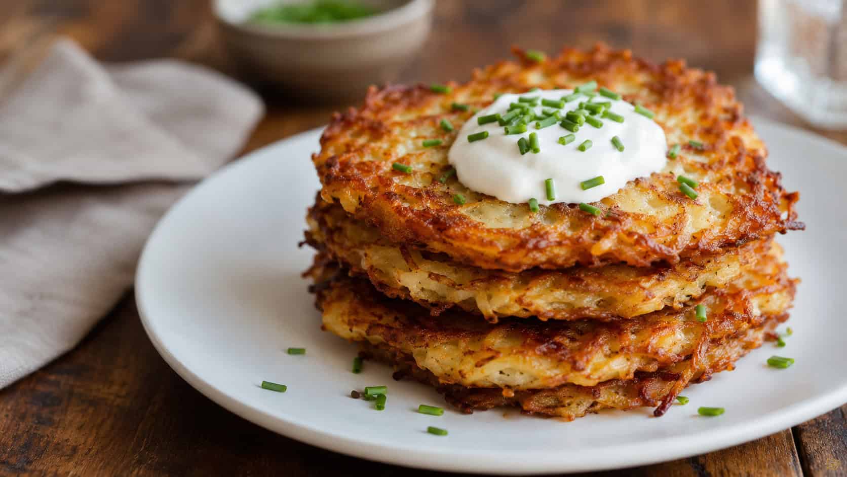 golden crispy potato pancakes in a wide horizontal shot, stacked with a dollop of sour cream and chives on top, on a white plate over a wooden table, warm natural light, shallow depth of field, landscape orientation