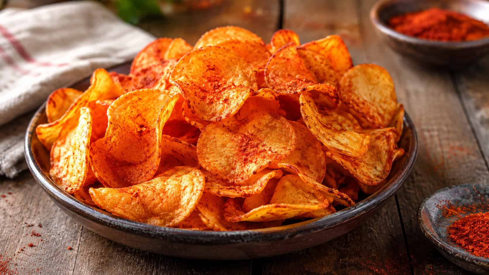 a pile of crispy paprika potato chips in a wide horizontal shot, reddish-gold and glistening, served in a shallow bowl on a rustic table with a sprinkle of paprika, bright natural light, shallow depth of field, landscape orientation