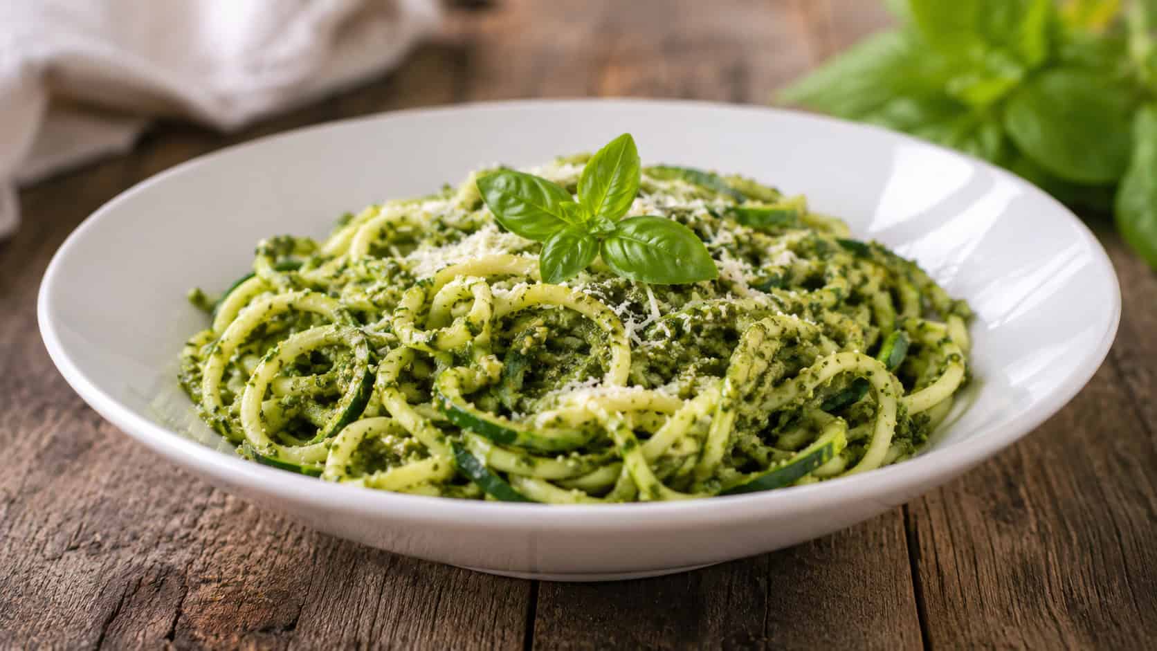a wide bowl of spiralized zucchini noodles glistening with green basil pesto and grated parmesan in a wide horizontal shot, shallow white bowl on a rustic wooden table with a sprig of basil, natural light, shallow depth of field, landscape orientation