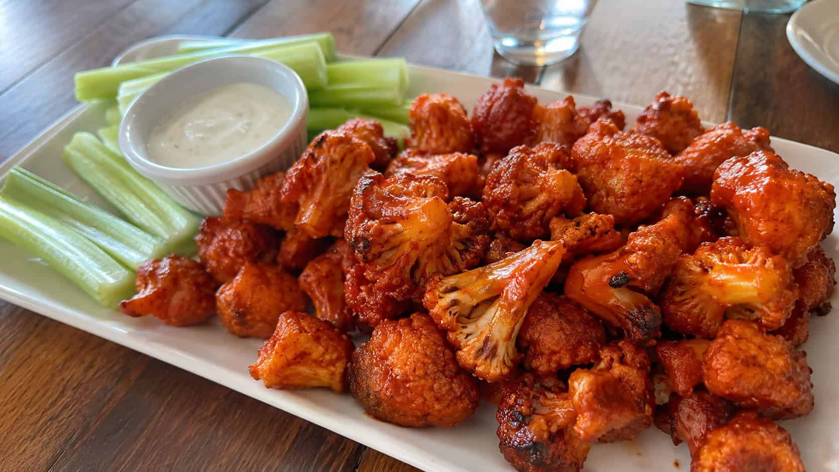 crispy roasted cauliflower bites coated in glossy red buffalo sauce in a wide horizontal shot, piled on a platter with celery sticks and a ramekin of ranch, natural light, shallow depth of field, landscape orientation