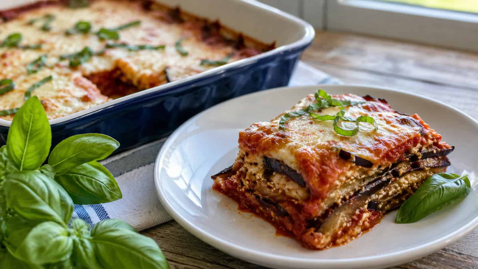a slice of eggplant lasagna with layers of cheese and marinara in a wide horizontal shot, plated beside the baking dish with fresh basil, natural light, shallow depth of field, landscape orientation