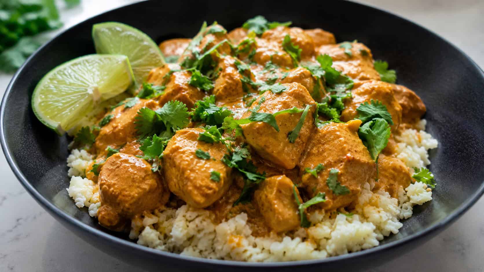 creamy orange coconut curry chicken in a wide horizontal shot, in a dark bowl over cauliflower rice with lime and cilantro garnish, natural light, shallow depth of field, landscape orientation