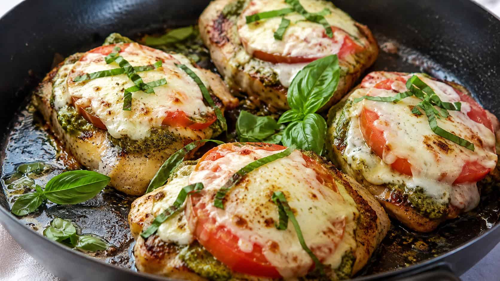 seared chicken breasts topped with green pesto, tomato, and melted mozzarella in a wide horizontal shot, in a skillet with fresh basil leaves, natural light, shallow depth of field, landscape orientation