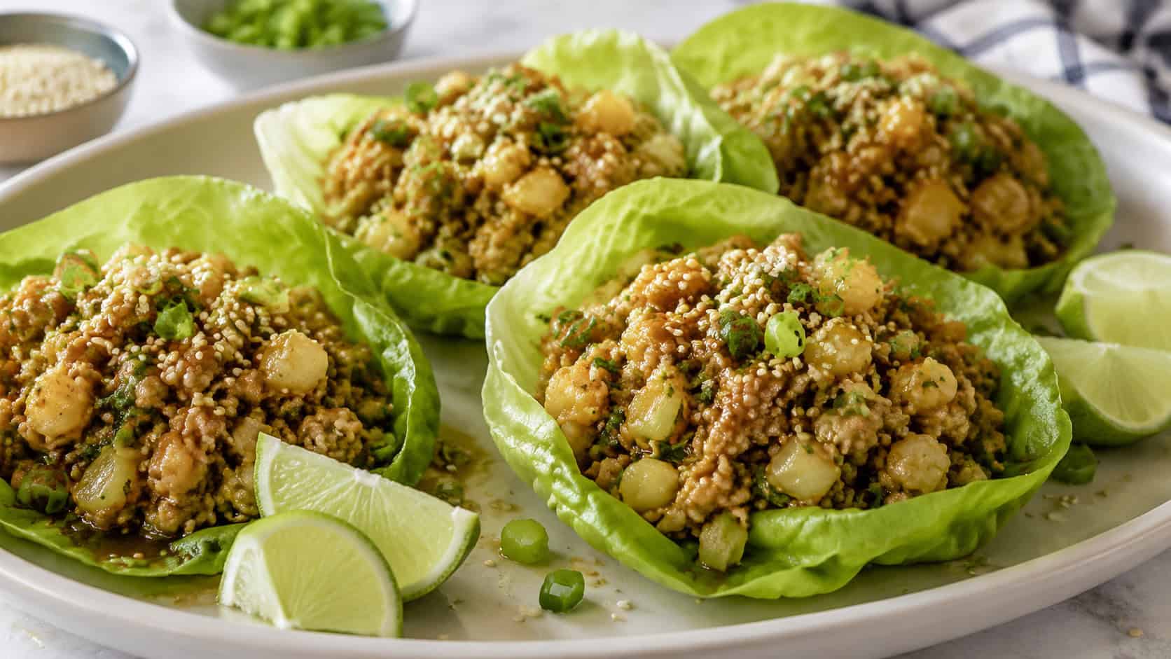 crisp butter lettuce cups filled with glossy seasoned ground chicken and water chestnuts in a wide horizontal shot, on a light platter with lime wedges and sesame seeds, natural light, shallow depth of field, landscape orientation