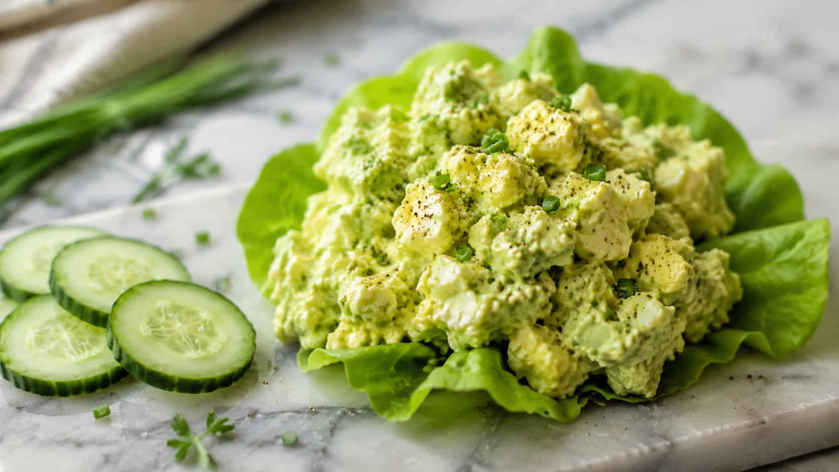 creamy green avocado egg salad mounded on crisp lettuce leaves in a wide horizontal shot, on a marble board with cucumber slices and chives, natural light, shallow depth of field, landscape orientation