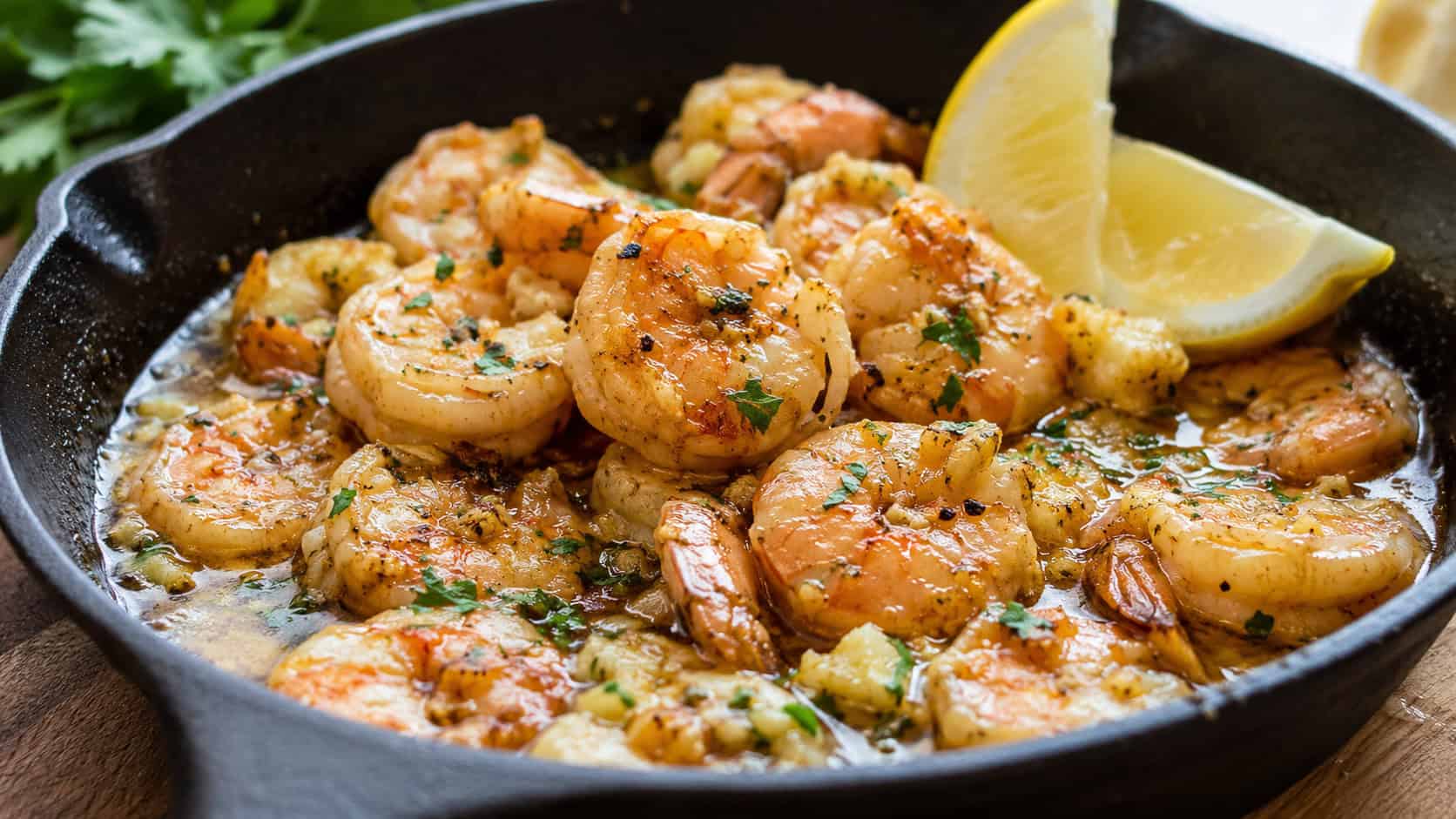 plump pink shrimp glistening in golden garlic butter sauce in a wide horizontal shot, in a cast iron skillet with lemon wedges and parsley, natural light, shallow depth of field, landscape orientation