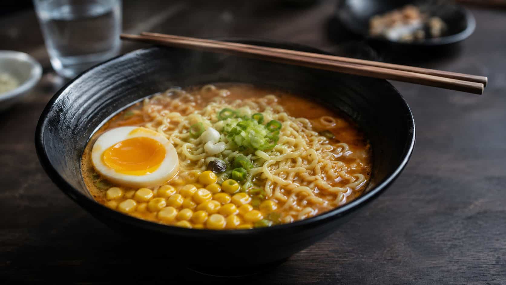 a steaming bowl of ramen in a wide horizontal shot, curly noodles in golden broth topped with a soft egg and green onions, chopsticks resting on the rim, set on a dark slate surface, moody warm light, shallow depth of field, landscape orientation