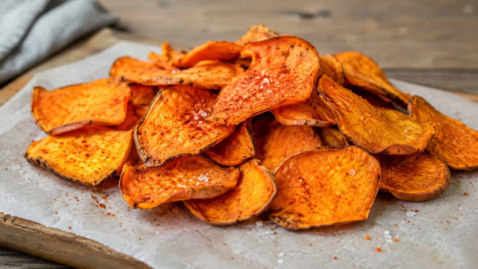a pile of crispy orange sweet potato chips in a wide horizontal shot, scattered across a parchment-lined board with a small dish of flaky salt, a striped towel in the background, bright natural light, shallow depth of field, landscape orientation