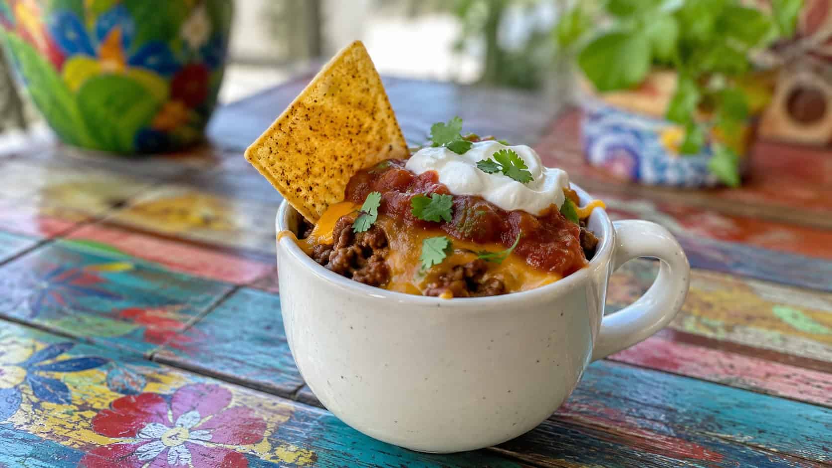 a taco-loaded mug in a wide horizontal shot, layers of seasoned beef, melted cheese, salsa, and crushed chips spilling over the rim, set on a colorful woven placemat, bright natural light, shallow depth of field, landscape orientation