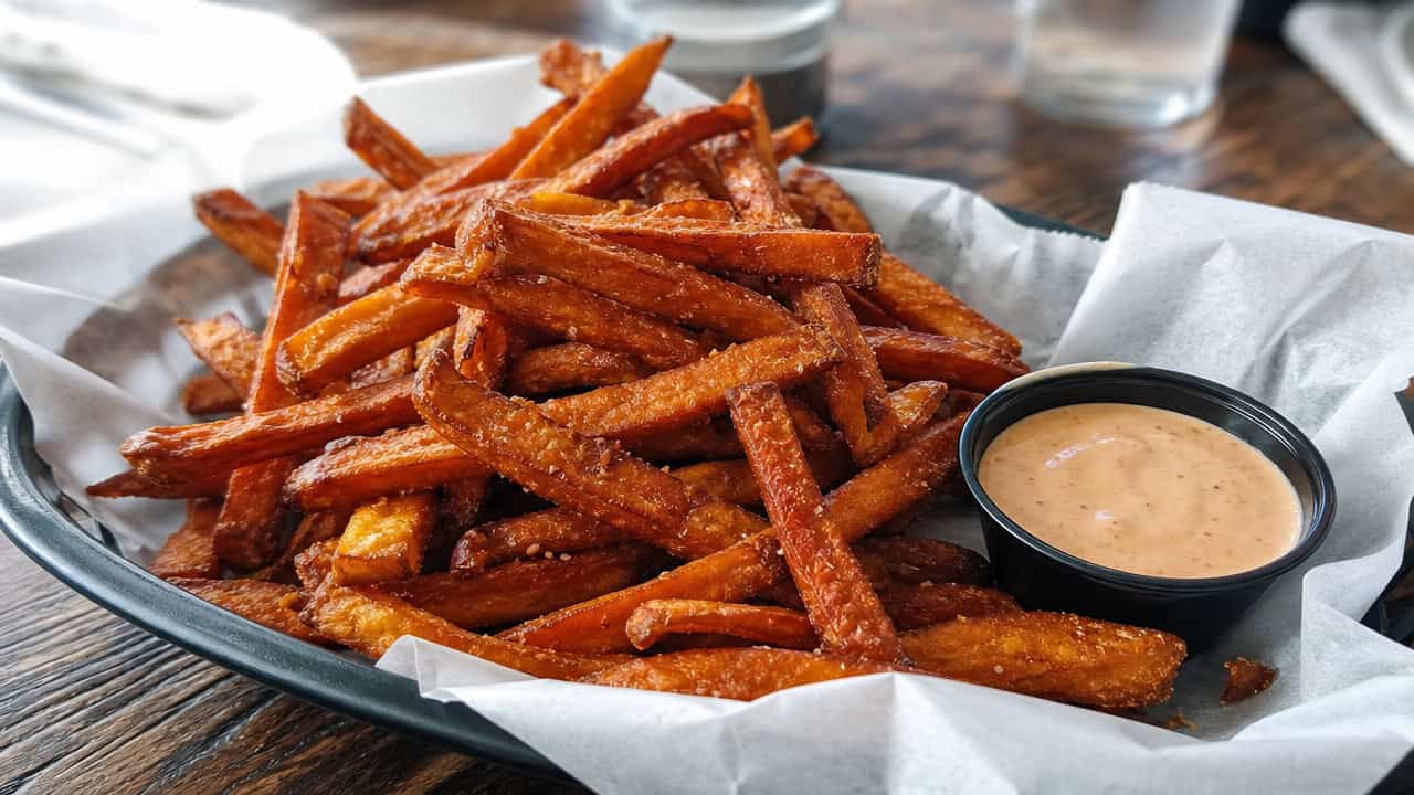 a pile of crispy sweet potato fries in a wide horizontal shot, served in a paper-lined basket with a dish of chipotle dipping sauce on a wooden table, natural light, shallow depth of field, landscape orientation