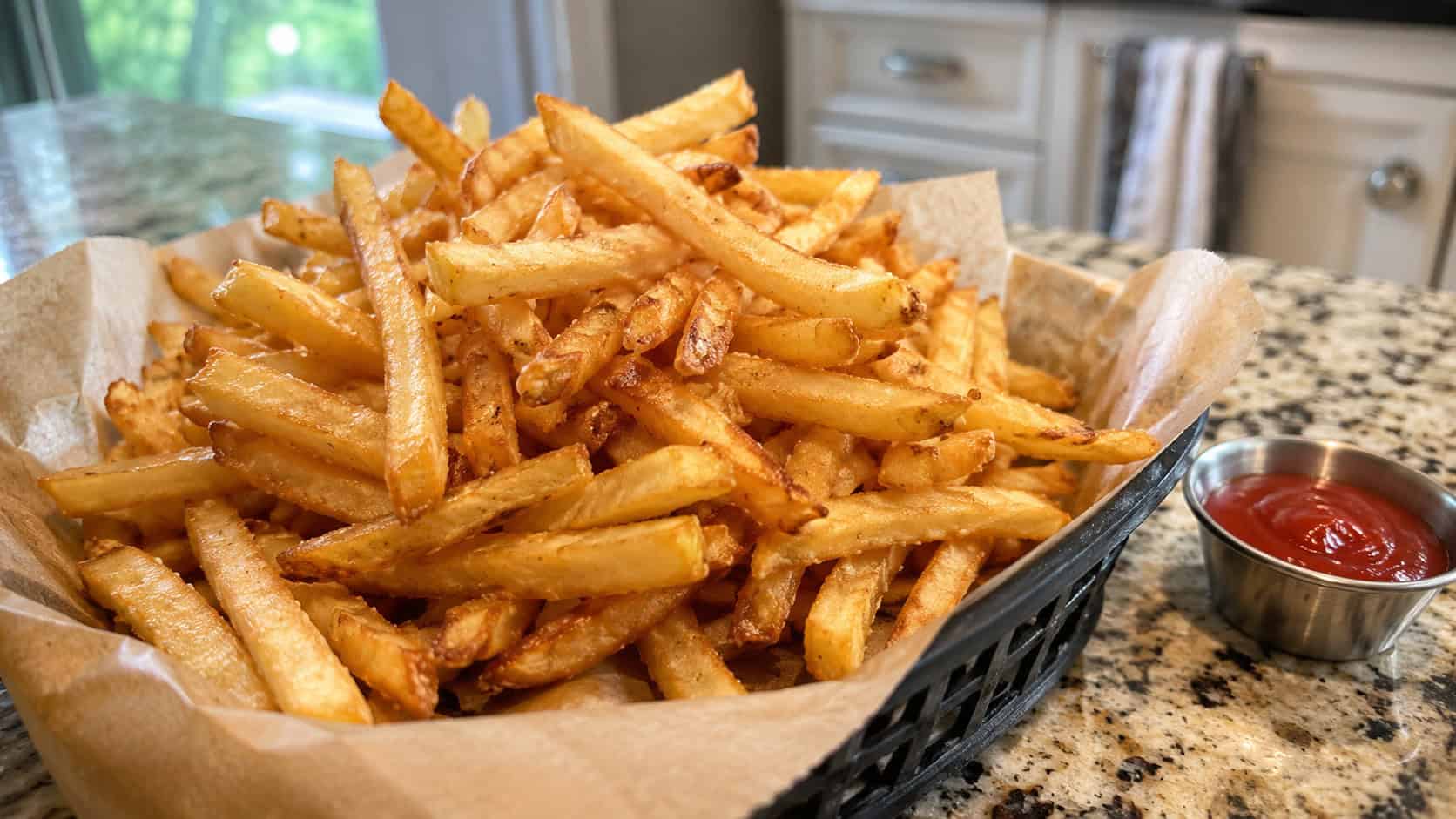 a generous heap of golden crispy french fries in a wide horizontal shot, spilling out of a paper-lined basket with a small dish of ketchup beside it on a kitchen counter, natural light, shallow depth of field, landscape orientation