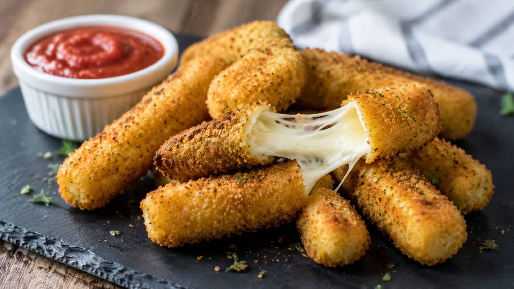 golden breaded mozzarella sticks with a gooey cheese pull in a wide horizontal shot, arranged on a slate board with a ramekin of marinara, natural light, shallow depth of field, landscape orientation