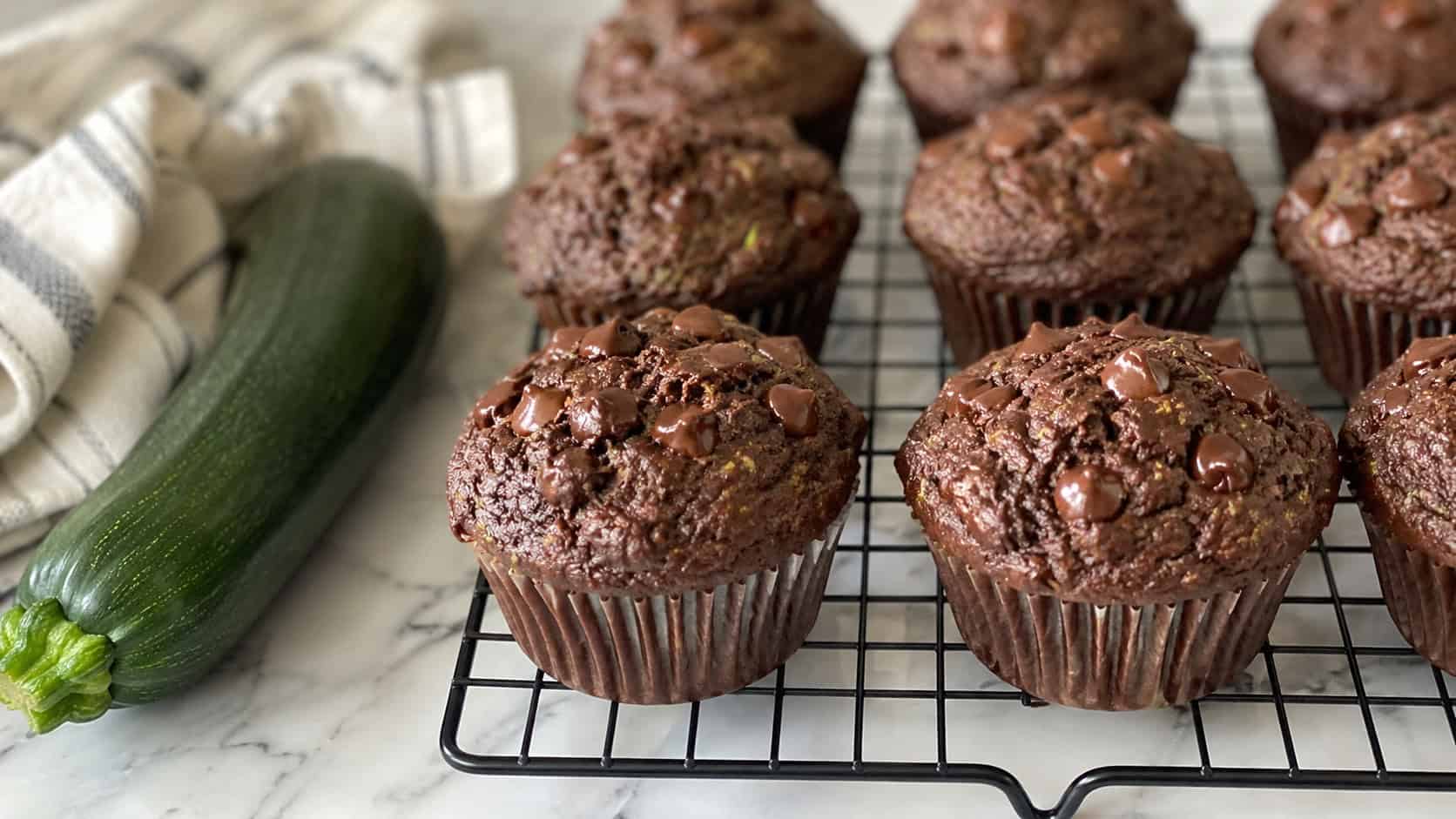 dark chocolate muffins with melty chocolate chips and faint green zucchini flecks in a wide horizontal shot, on a cooling rack beside a small zucchini on a marble counter, natural light, shallow depth of field, landscape orientation