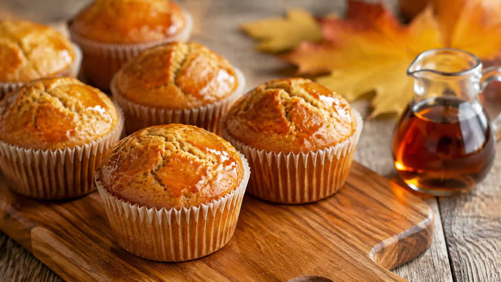 golden maple muffins with glossy syrup-brushed domed tops in a wide horizontal shot, on a wooden board beside a small jug of maple syrup on a warm rustic table, natural light, shallow depth of field, landscape orientation