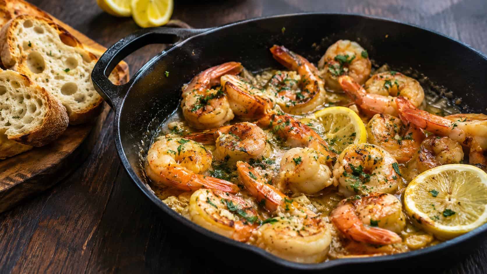 plump shrimp glistening in a buttery garlic sauce with lemon and parsley in a wide horizontal shot, served in a cast iron skillet with crusty bread alongside on a dark wooden table, natural light, shallow depth of field, landscape orientation