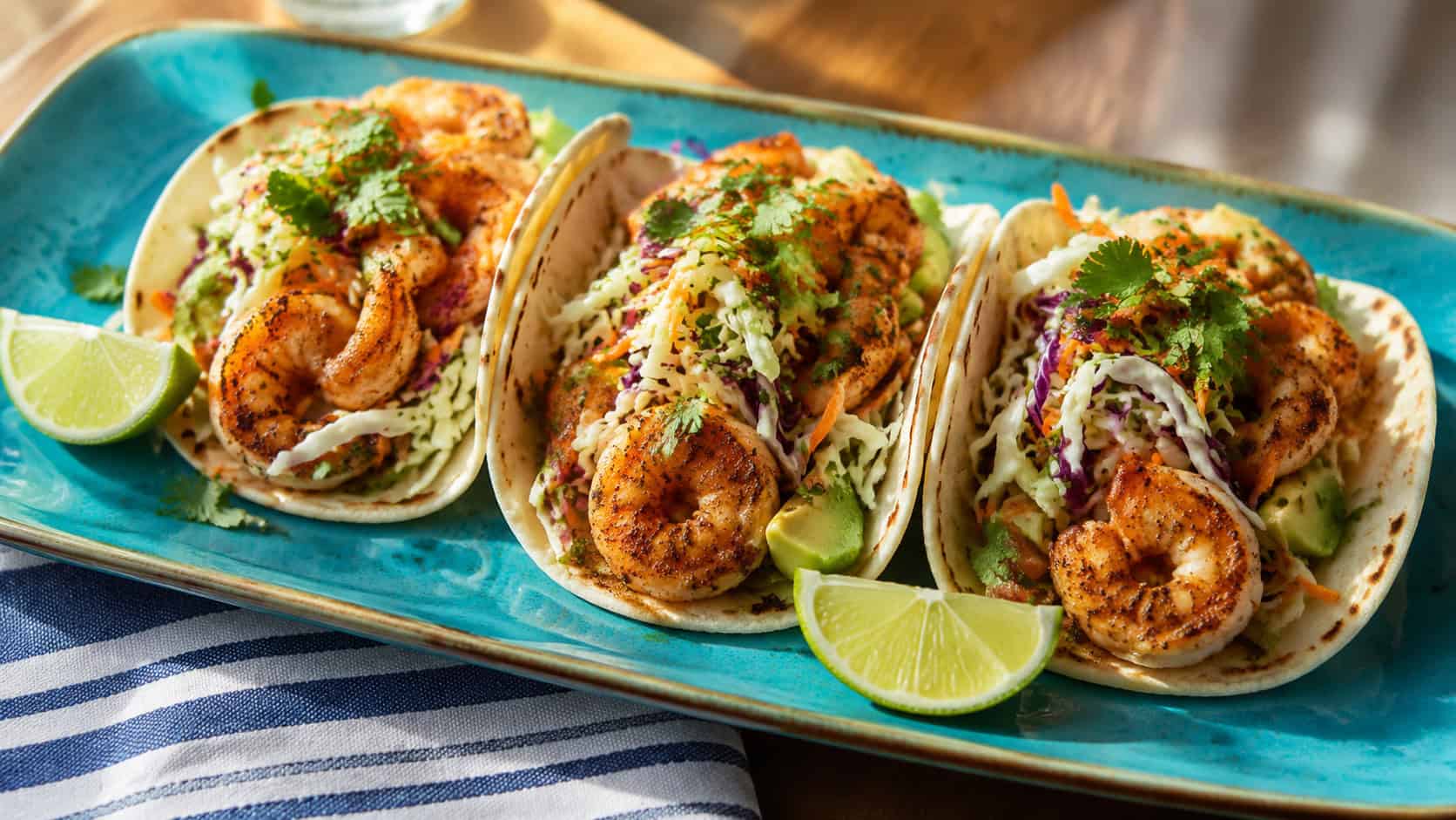 three shrimp tacos lined up on a wide horizontal platter, topped with crunchy slaw, cilantro and lime wedges, on a bright blue plate with a striped napkin on a sunny table, natural light, shallow depth of field, landscape orientation