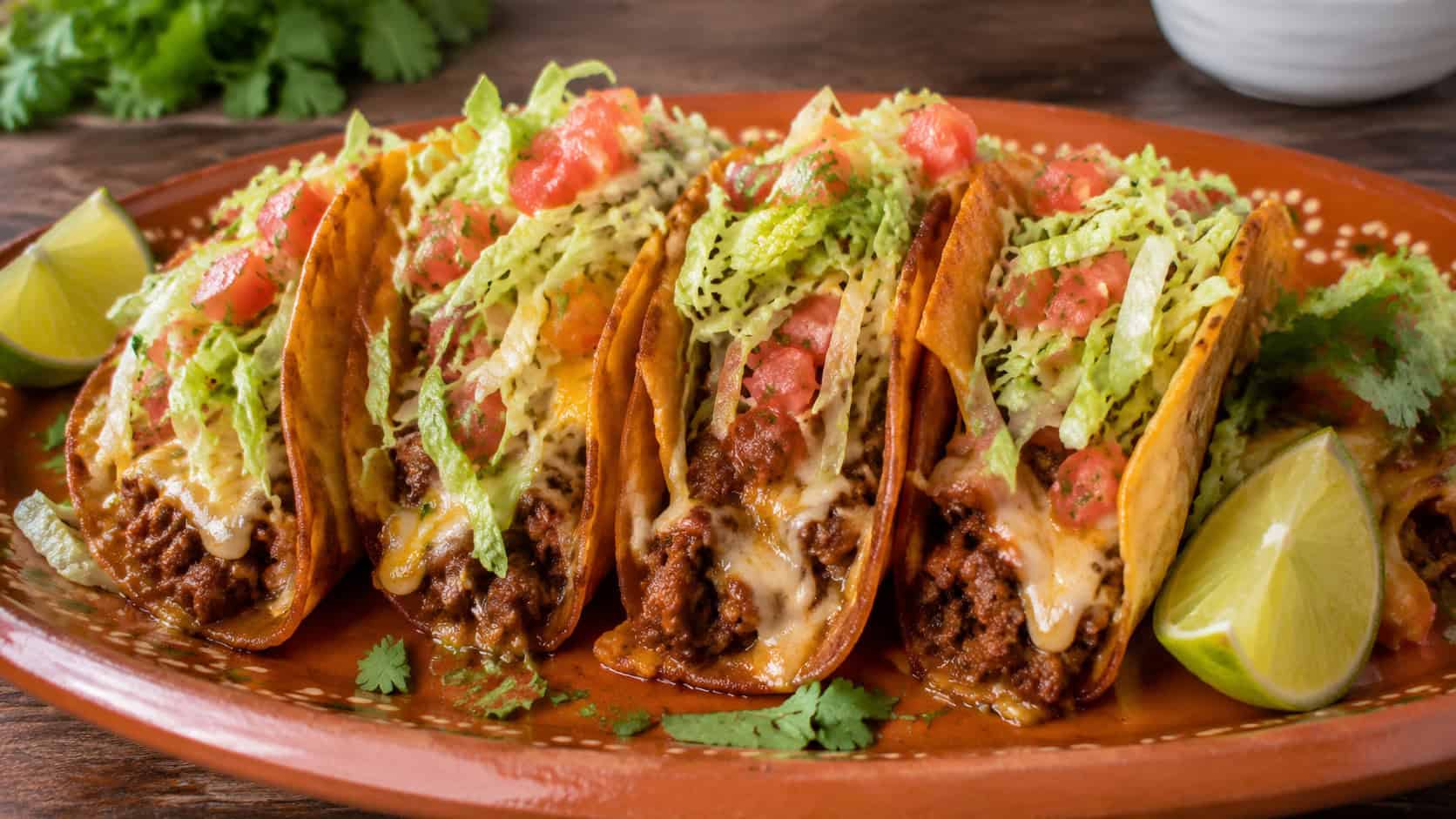 a row of crisp beef tacos overflowing with melted cheese, lettuce and diced tomato in a wide horizontal shot, on a warm terracotta platter with lime and cilantro scattered around on a wooden table, natural light, shallow depth of field, landscape orientation