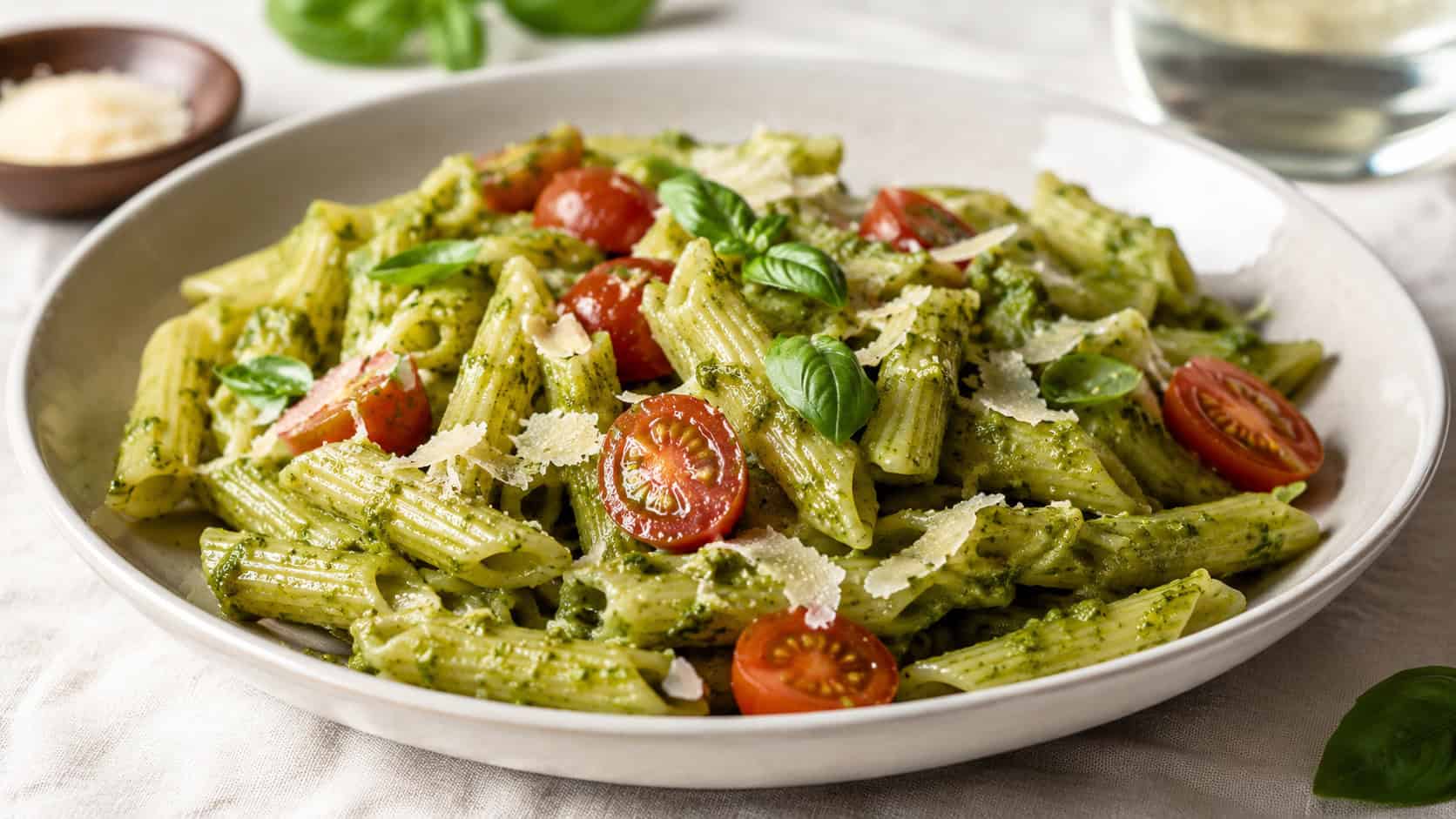 vibrant green pesto-coated penne studded with halved cherry tomatoes in a wide horizontal shot, served in a wide shallow bowl with shaved Parmesan and basil on a light linen tablecloth, natural light, shallow depth of field, landscape orientation