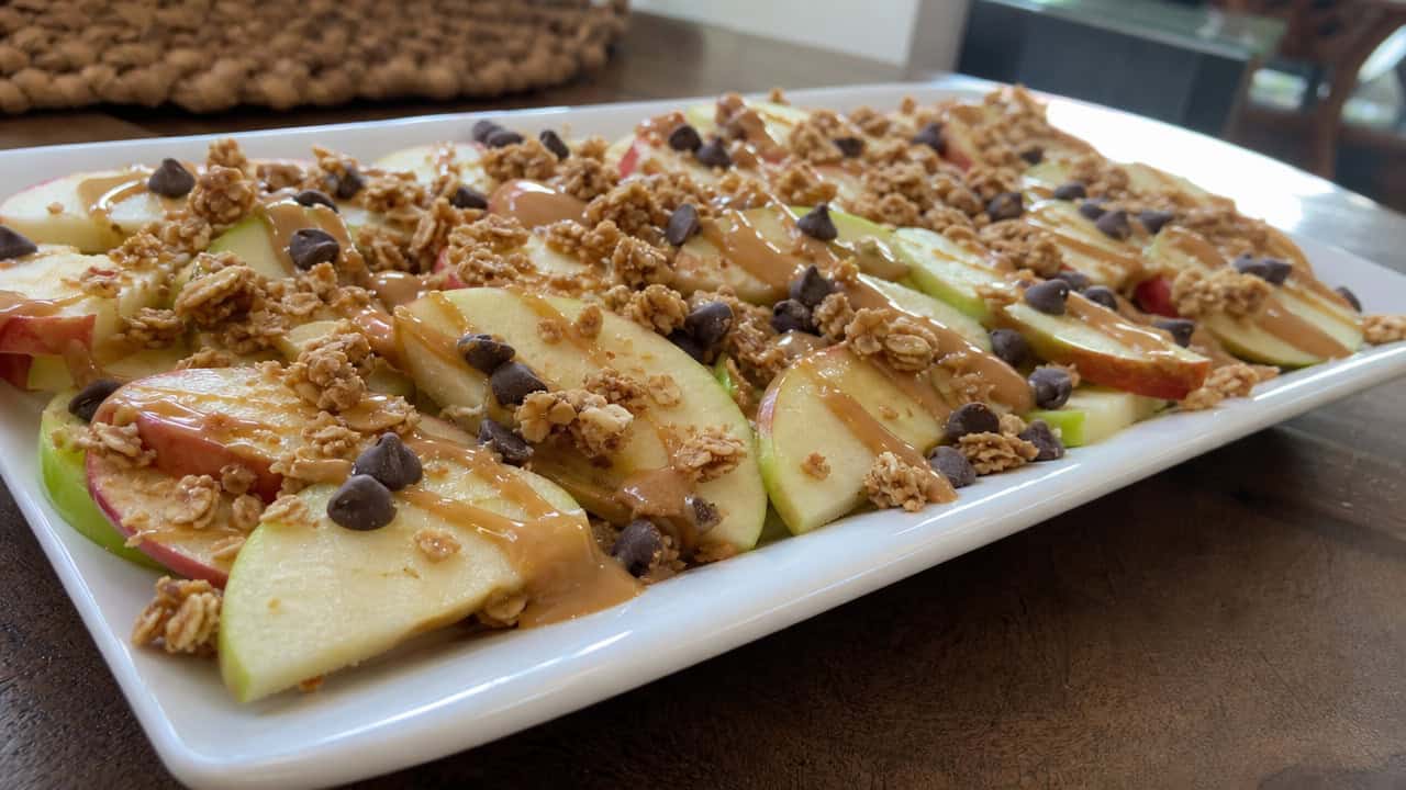 apple nachos with thin apple slices drizzled in peanut butter, chocolate chips, and granola spread across a wide platter on a table, natural light, shallow depth of field, landscape orientation