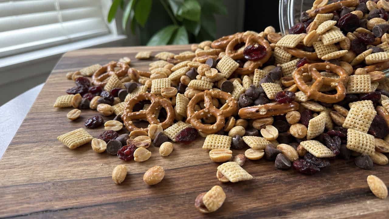 homemade trail mix with pretzels, cereal, peanuts, chocolate chips, and dried cranberries spilling across a wide wooden board, natural light, shallow depth of field, landscape orientation