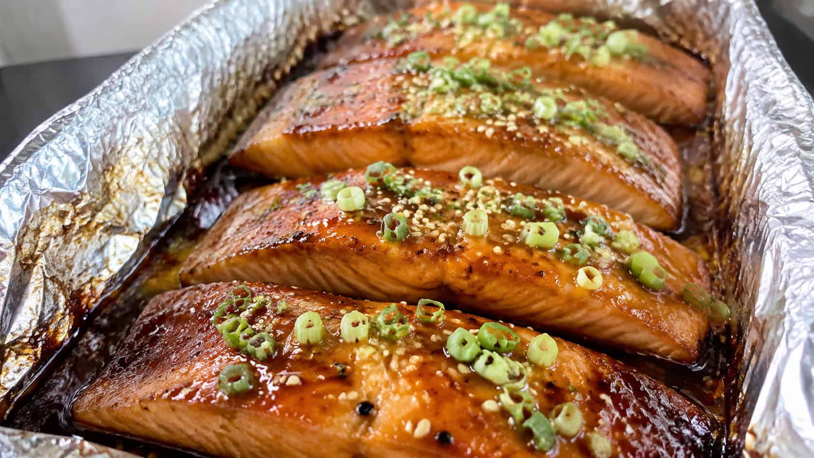honey soy baked salmon in a wide horizontal shot, glossy glazed fillets topped with green onion and sesame seeds in a foil-lined dish, natural light, shallow depth of field, landscape orientation