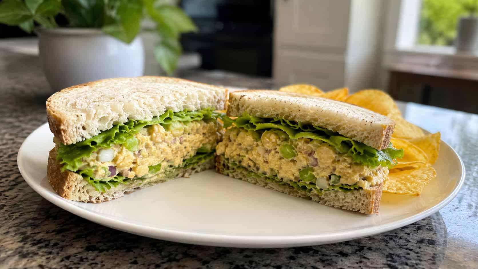 a wide chickpea salad sandwich cut in half showing the creamy mashed chickpea filling and crisp lettuce, on a plate with chips beside it, on a kitchen counter, natural light, shallow depth of field, landscape orientation
