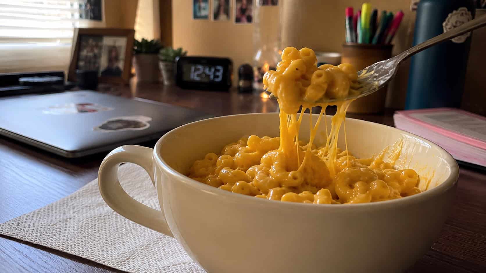 a wide creamy mug of macaroni and cheese with a glossy cheddar sauce, a fork lifting a cheesy bite, on a dorm desk with a napkin, warm natural light, shallow depth of field, landscape orientation