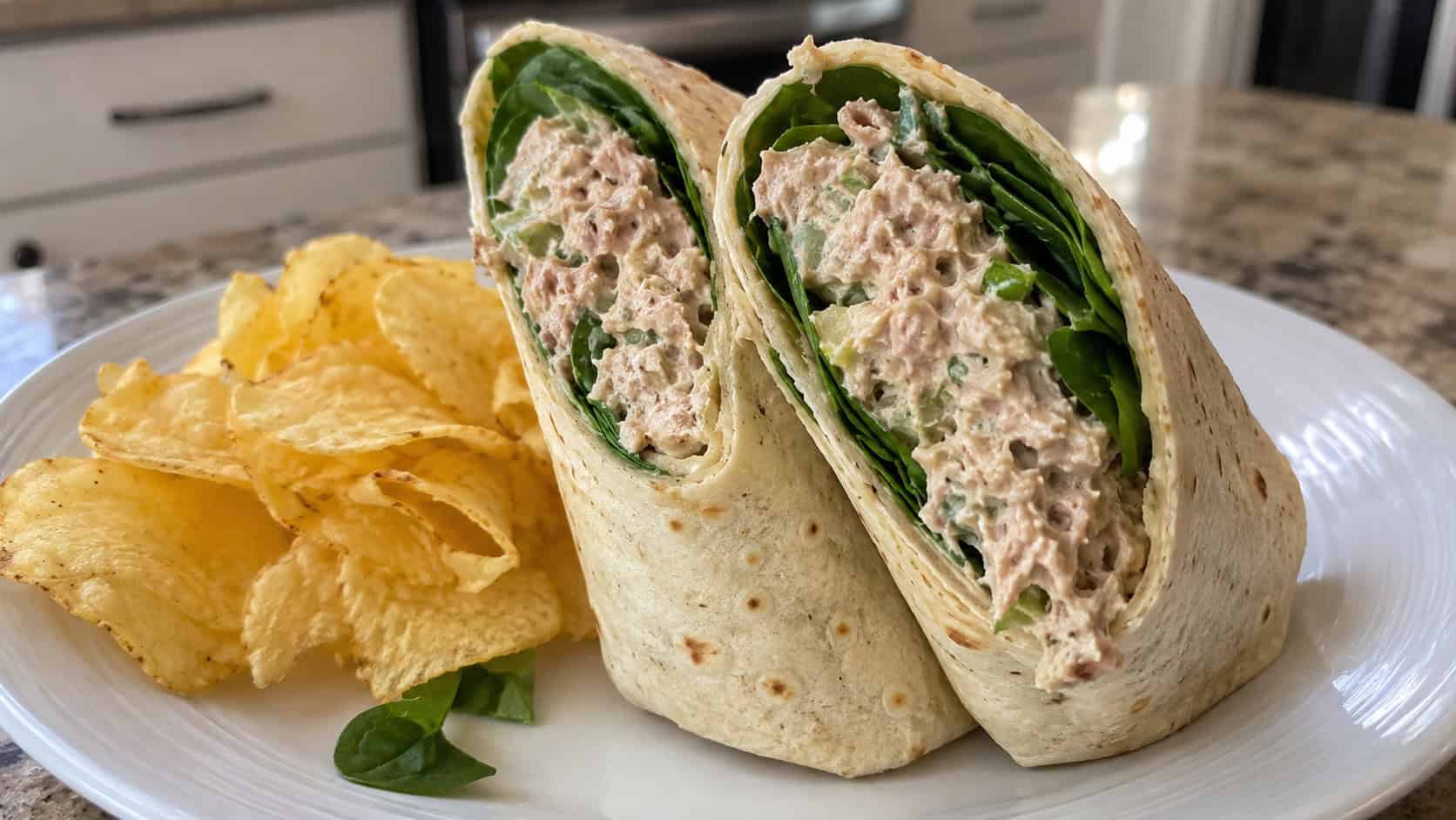 a wide tuna salad wrap cut in half on the diagonal showing creamy tuna and fresh spinach, standing upright on a plate with chips beside it, on a counter, natural light, shallow depth of field, landscape orientation