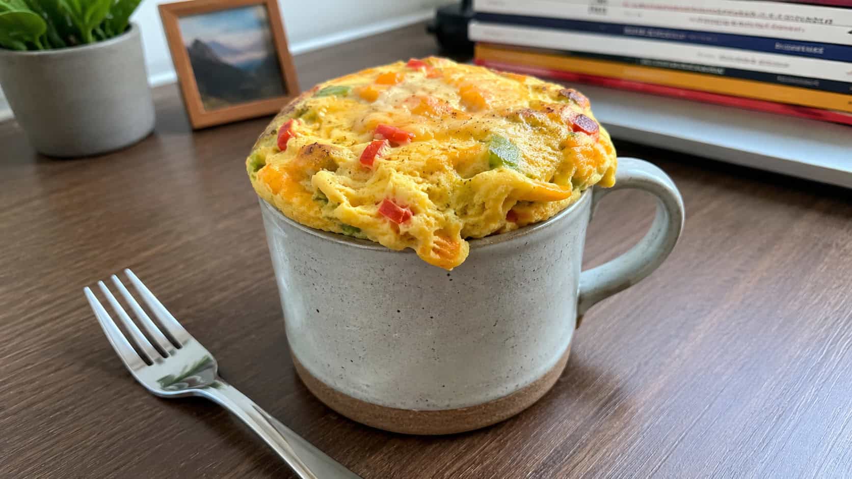 a fluffy mug omelette puffing over the rim of a ceramic mug, flecked with melted cheese and colorful peppers, on a wide desk with a fork beside it, bright natural light, shallow depth of field, landscape orientation