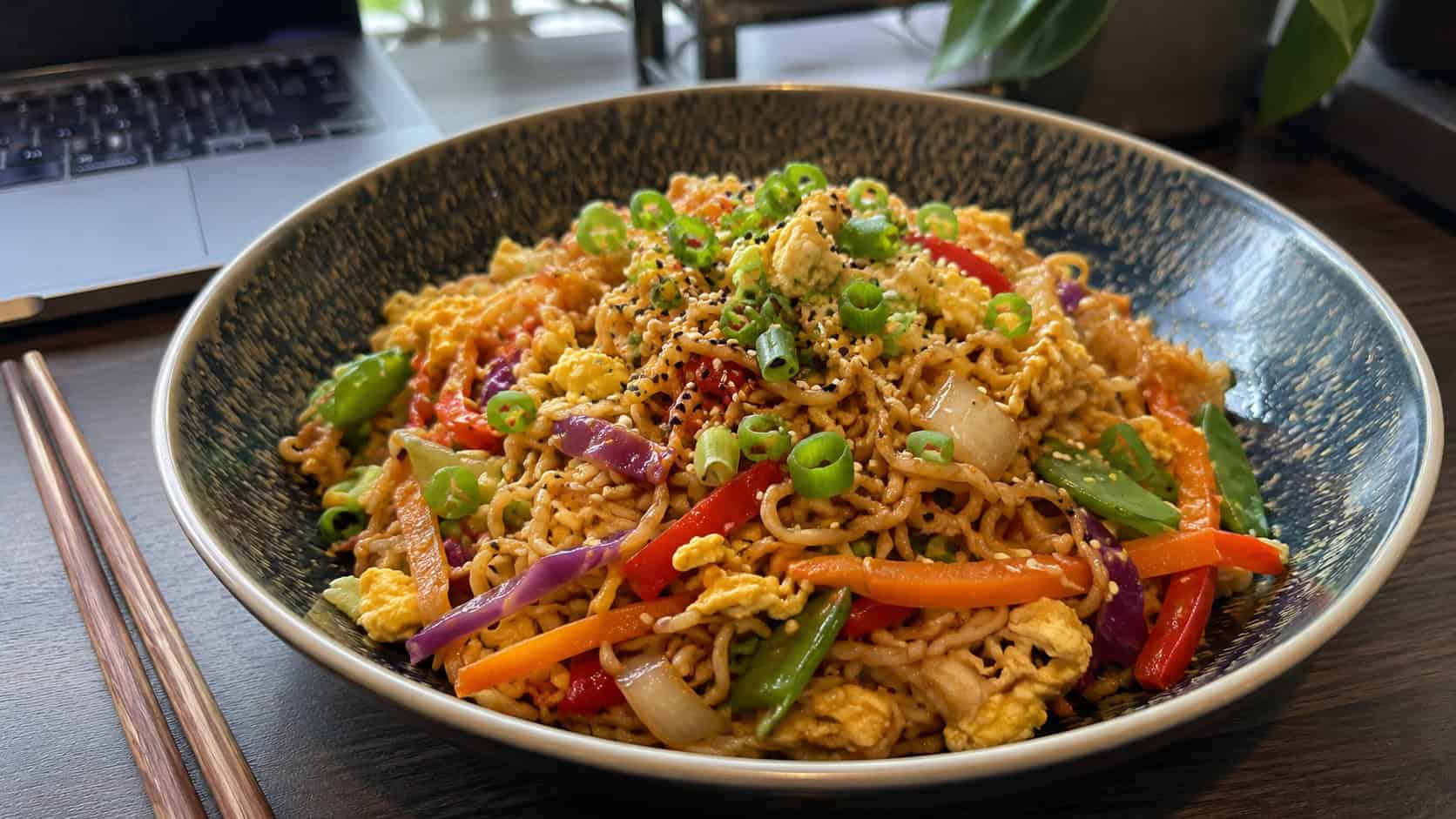 a wide bowl of ramen stir-fry with glossy noodles, colorful vegetables, scrambled egg, and sliced green onion, sesame seeds on top, chopsticks resting beside, on a desk, natural light, shallow depth of field, landscape orientation