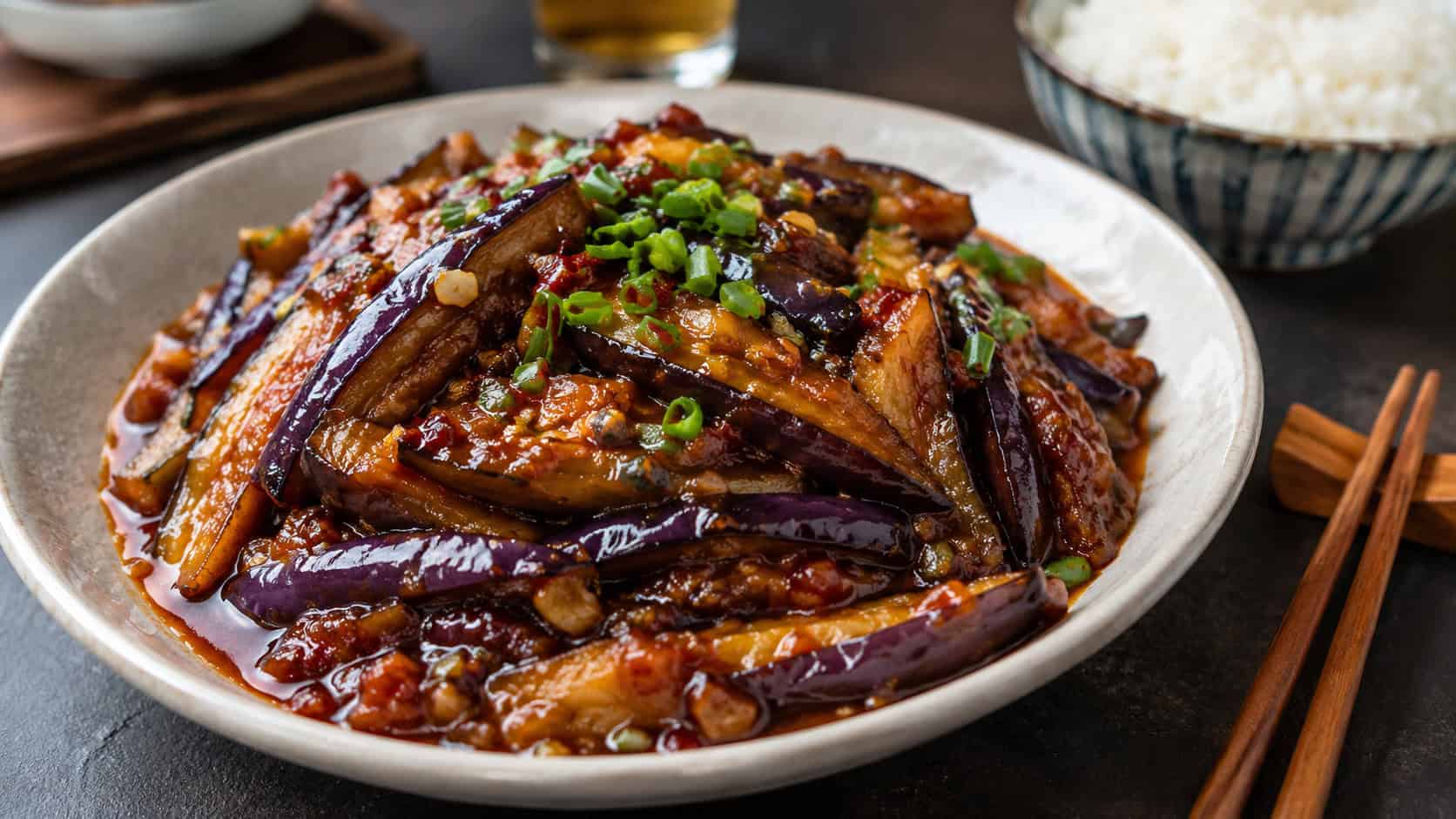 glossy braised Szechuan eggplant in a wide horizontal shot, deep red sauce clinging to tender strips, scattered scallions and chili flakes, chopsticks and steamed rice nearby on a dark table, warm natural light, shallow depth of field, landscape orientation