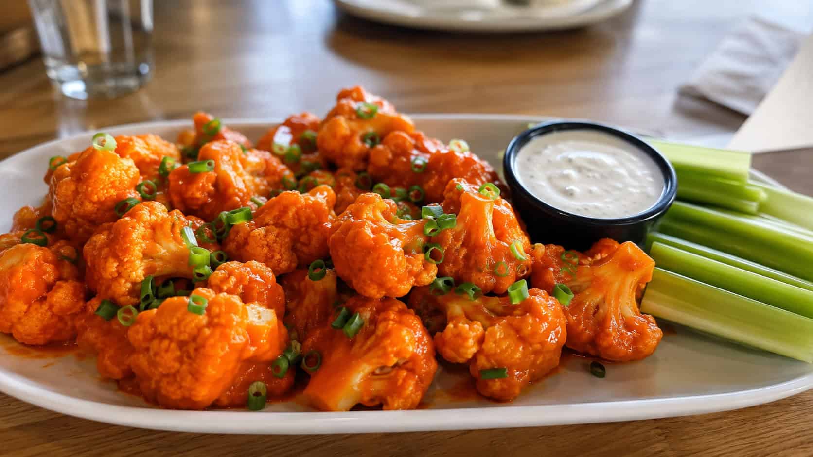a wide platter of saucy orange buffalo cauliflower bites in a horizontal shot, glistening and dusted with green onion, a small bowl of ranch and celery sticks alongside on a casual table, bright natural light, shallow depth of field, landscape orientation