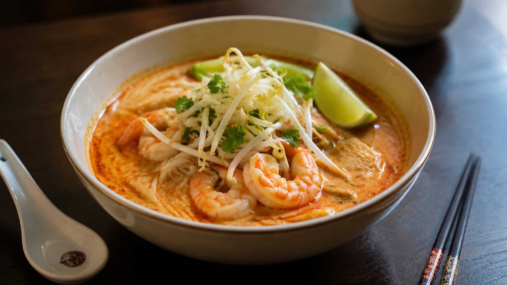 a wide bowl of red coconut curry laksa in a horizontal shot, noodles and pink shrimp peeking through the creamy orange broth, topped with bean sprouts and lime, a spoon and chopsticks beside it on a dark table, warm natural light, shallow depth of field, landscape orientation