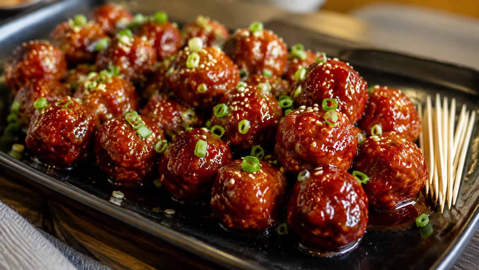 a wide platter of glossy sriracha honey glazed meatballs in a horizontal shot, deep red and shiny with sauce, sprinkled with scallions and sesame seeds, toothpicks tucked in beside them on a dark serving board, warm natural light, shallow depth of field, landscape orientation