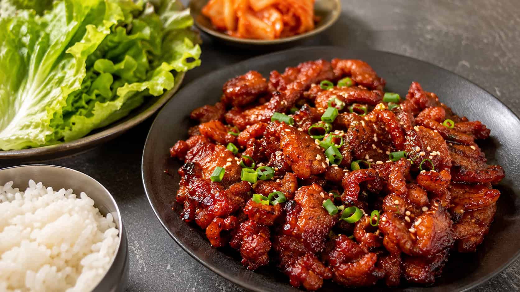 caramelized spicy Korean pork bulgogi in a wide horizontal shot, deep red and glistening with sauce, scattered scallions and sesame seeds, lettuce leaves and rice alongside on a dark stone table, warm natural light, shallow depth of field, landscape orientation