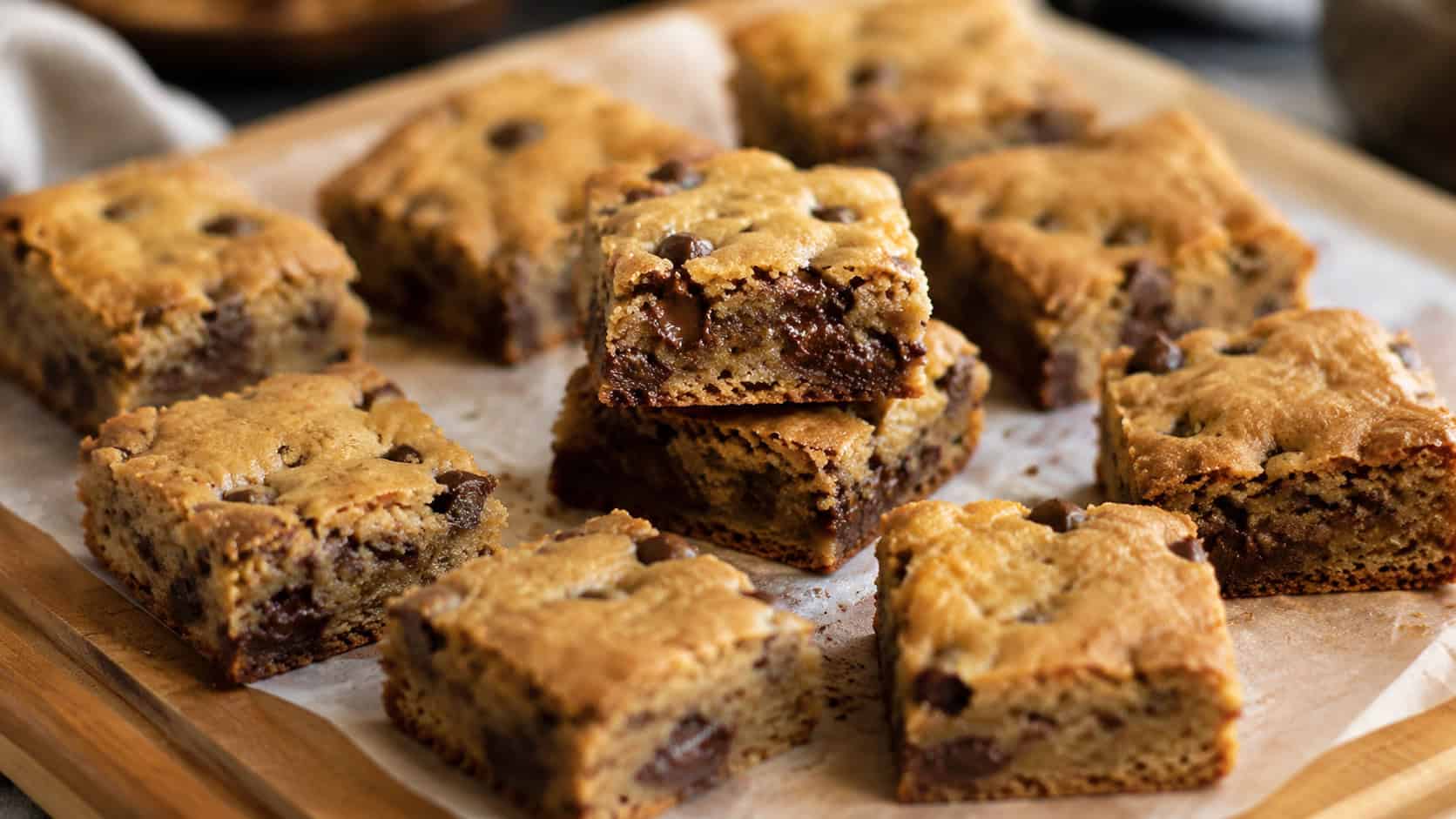 banana chocolate chip blondies cut into squares on parchment, melty chocolate chips throughout, a few stacked, on a wooden board, wide horizontal shot, warm natural light, shallow depth of field, landscape orientation