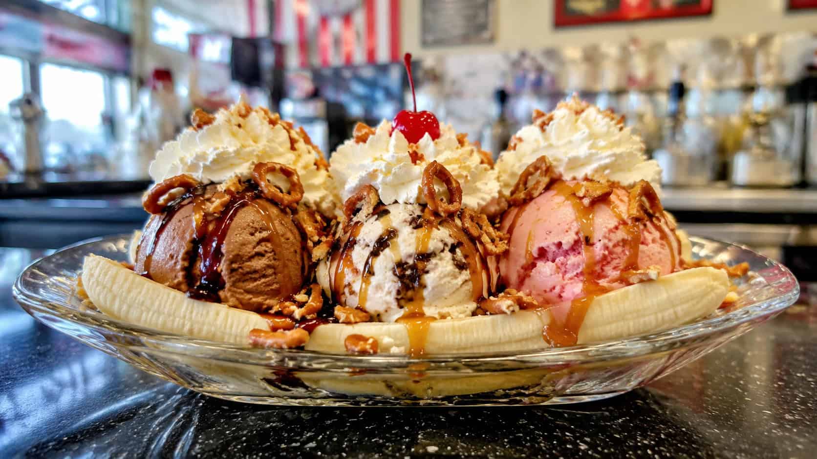 a loaded banana split in a long dish with three ice cream scoops, caramel and chocolate drizzle, crushed pretzels, and whipped cream, on a diner counter, wide horizontal shot, bright light, shallow depth of field, landscape orientation