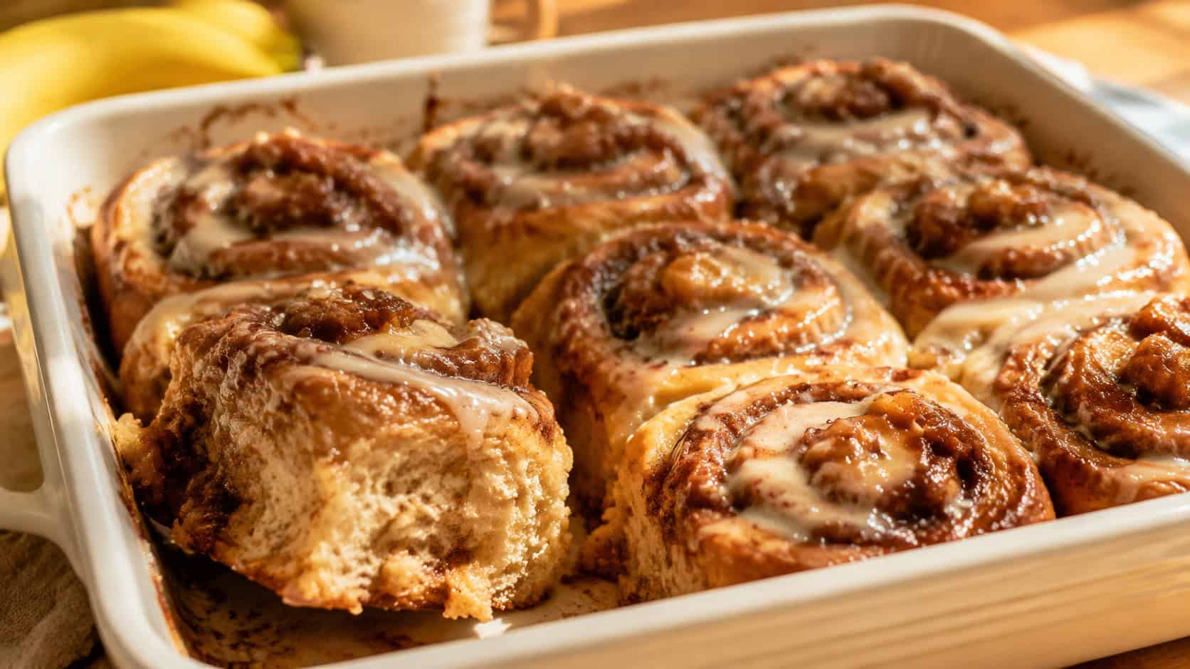 gooey banana cinnamon rolls in a baking dish drizzled with white glaze, swirls of cinnamon visible, one roll pulled out, wide horizontal shot, warm morning light, shallow depth of field, landscape orientation