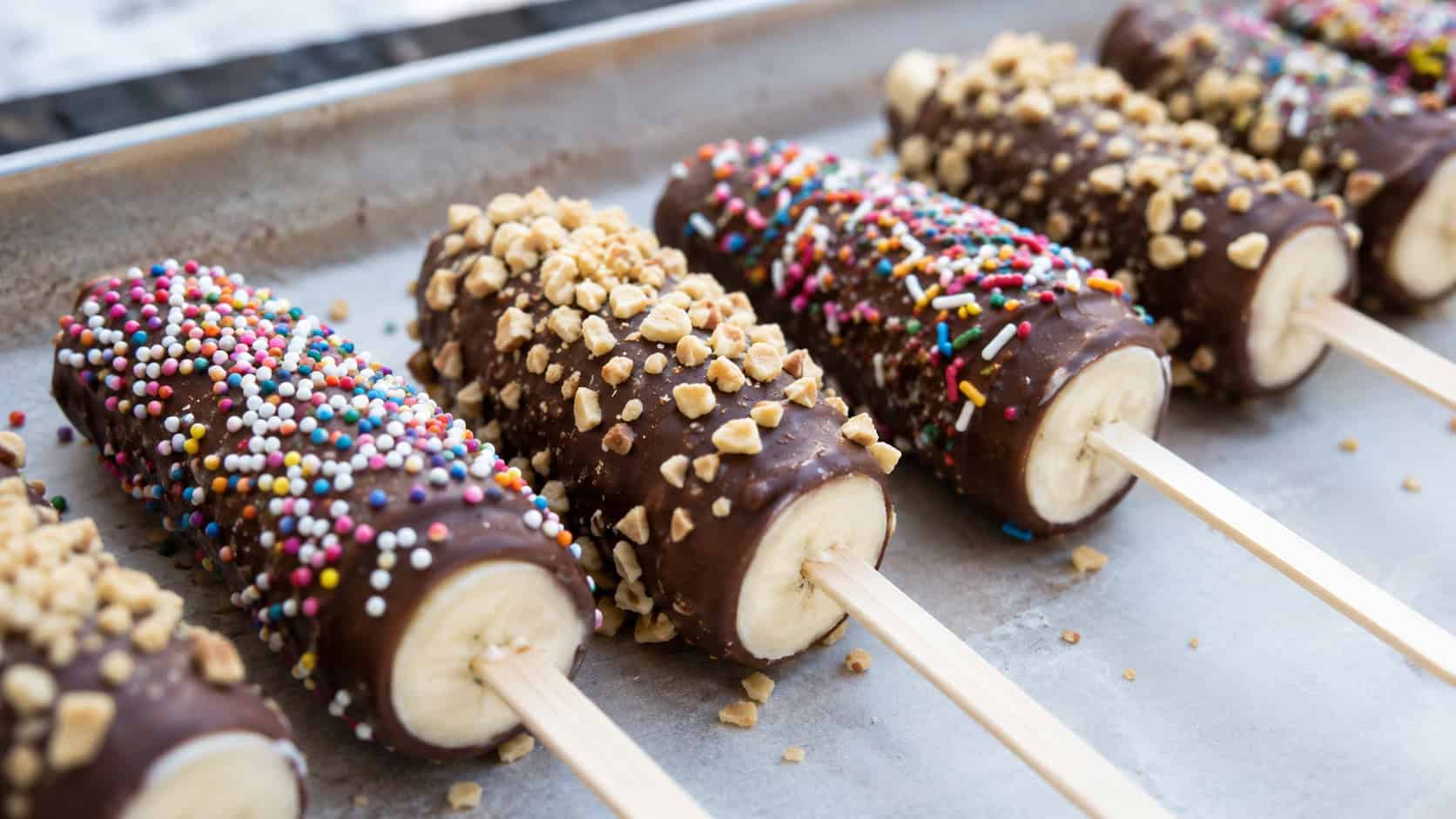 frozen chocolate-dipped banana pops on sticks covered in sprinkles and crushed peanuts, lined up on a parchment tray, wide horizontal composition, cool bright light, shallow depth of field, landscape orientation