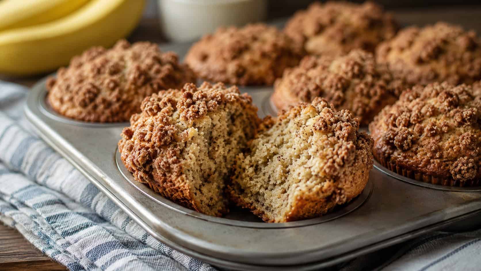 banana muffins with crumbly cinnamon streusel tops cooling in a muffin tin, one split open showing tender crumb, on a kitchen towel, wide horizontal composition, warm natural light, shallow depth of field, landscape orientation