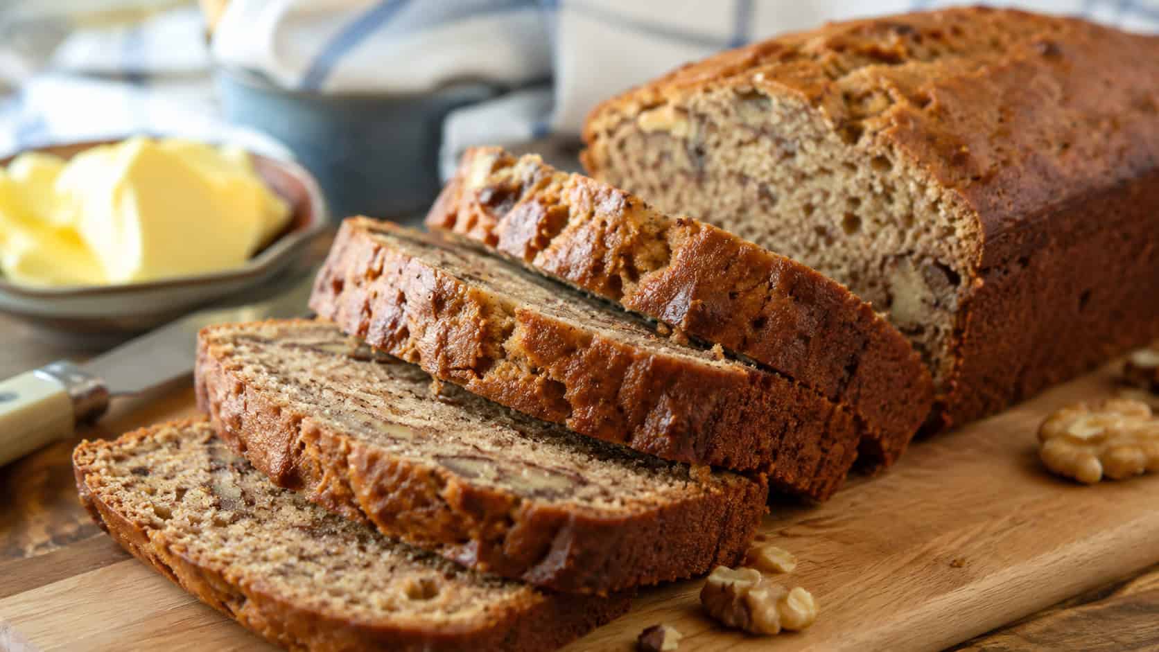 a sliced loaf of walnut banana bread on a wooden cutting board, a few slices fanned out showing moist crumb and walnuts, butter nearby, wide horizontal composition, warm natural light, shallow depth of field, landscape orientation