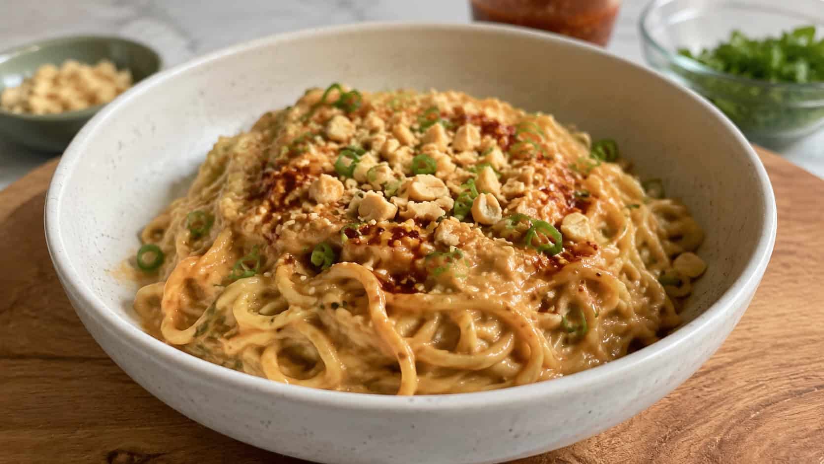 a wide bowl of peanut sauce ramen with creamy tan-coated noodles and chopped peanuts on top, on a wooden board, natural light, shallow depth of field, landscape orientation