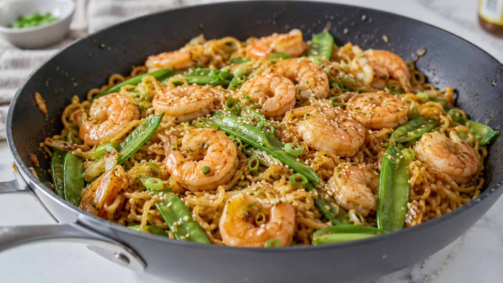 a wide skillet of garlic shrimp ramen stir-fry with glossy noodles, plump pink shrimp, bright green snap peas and a sprinkle of sesame seeds, on a light countertop, natural light, shallow depth of field, landscape orientation
