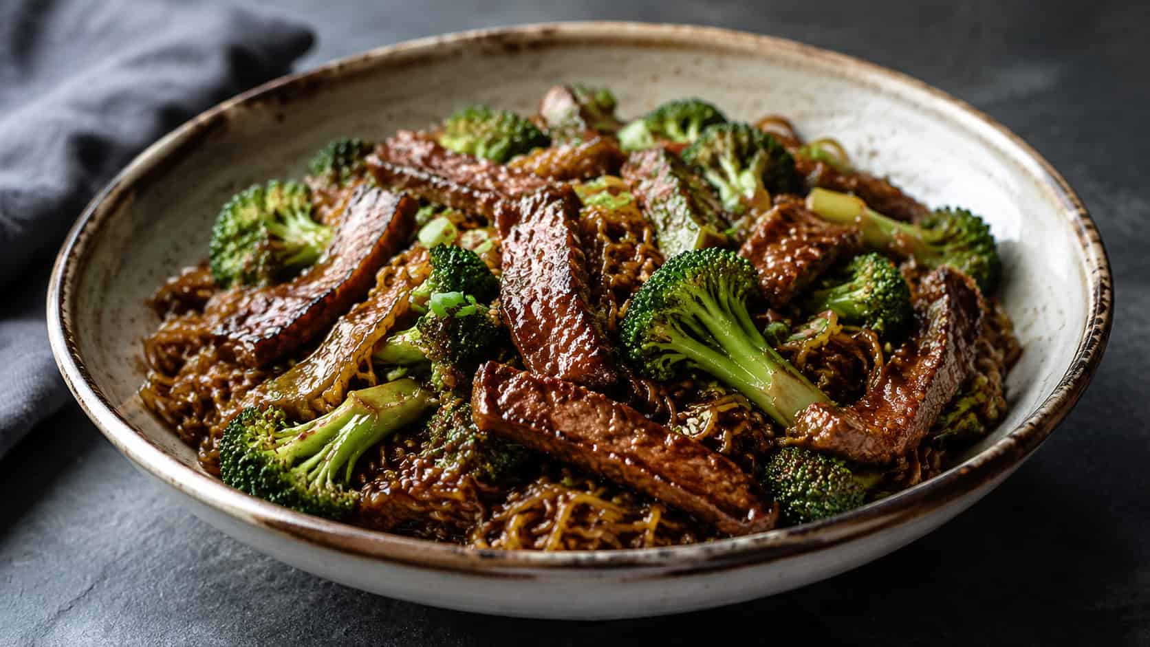 a wide bowl of beef and broccoli ramen with glossy dark-sauced noodles, strips of seared beef and bright green broccoli, on a slate surface, natural light, shallow depth of field, landscape orientation