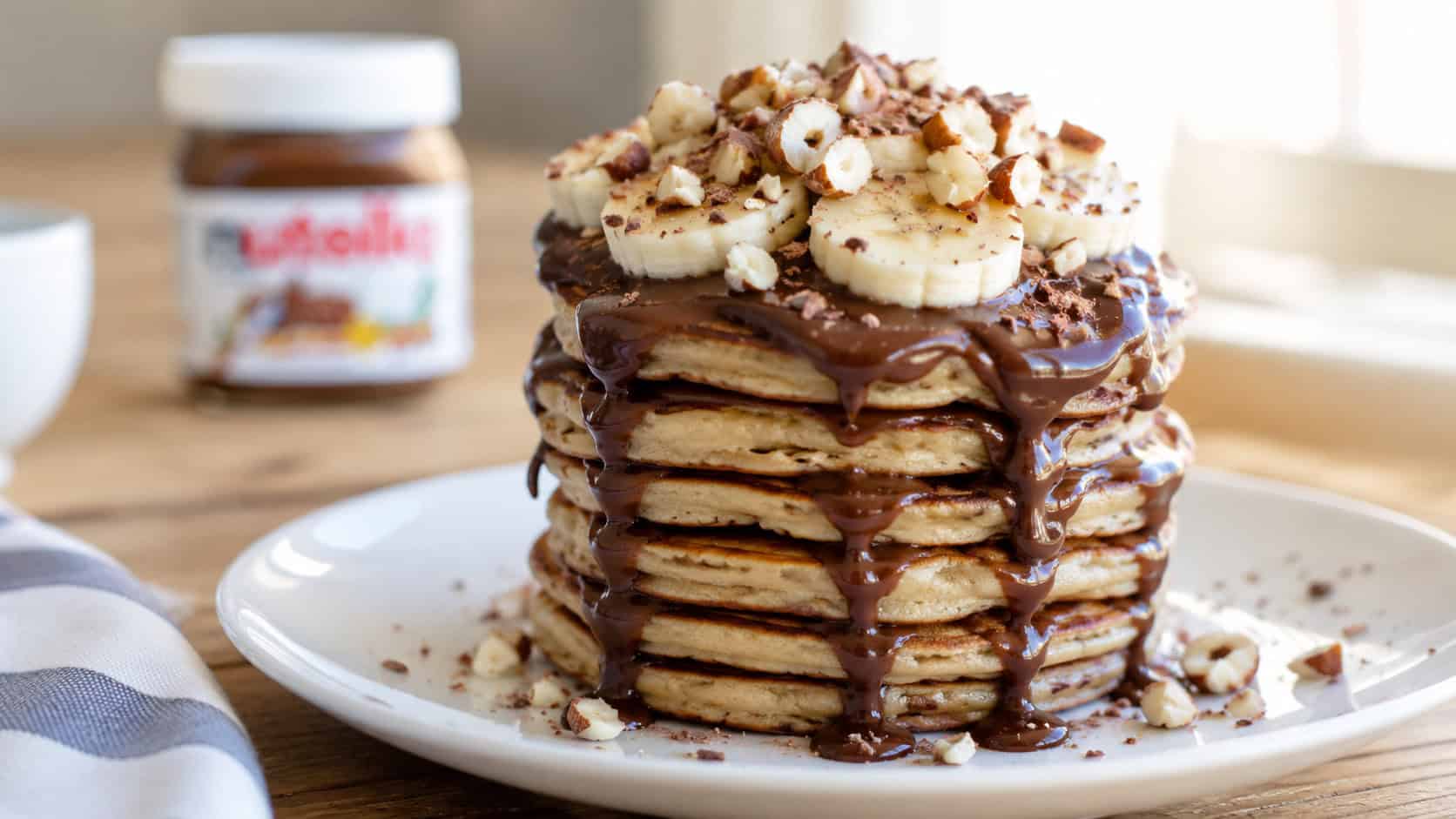 a tall stack of Nutella pancakes on a plate in a wide horizontal shot, chocolate hazelnut dripping down the sides, banana slices and chopped hazelnuts on top, bright breakfast light, shallow depth of field, landscape orientation