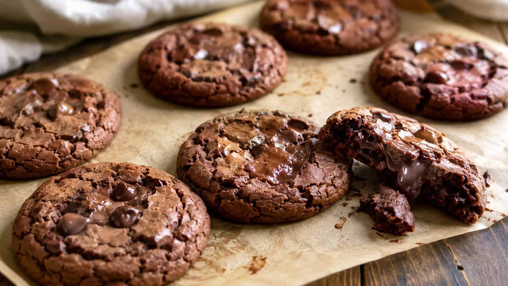 Nutella cookies cooling on parchment in a wide horizontal shot, crackly tops with melty chocolate chips, one cookie broken to show a chewy center, rustic wood surface, warm natural light, shallow depth of field, landscape orientation