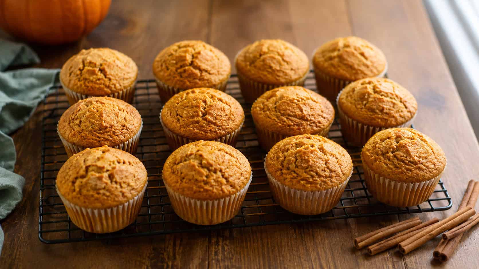 a dozen golden pumpkin spice muffins in paper liners cooling on a wire rack, shown in a wide horizontal shot on a warm wooden table with cinnamon sticks nearby, natural light, shallow depth of field, landscape orientation