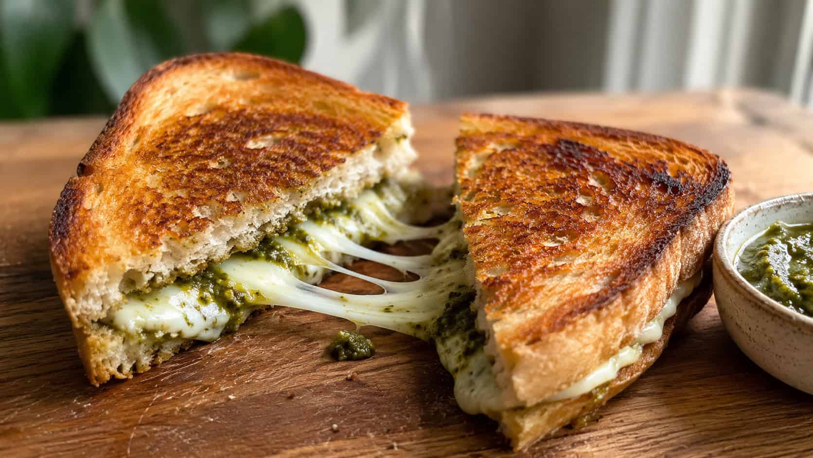 a golden grilled cheese cut in half with melty mozzarella and green pesto pulling apart, shown in a wide horizontal shot on a wooden board with a small bowl of pesto nearby, natural light, shallow depth of field, landscape orientation