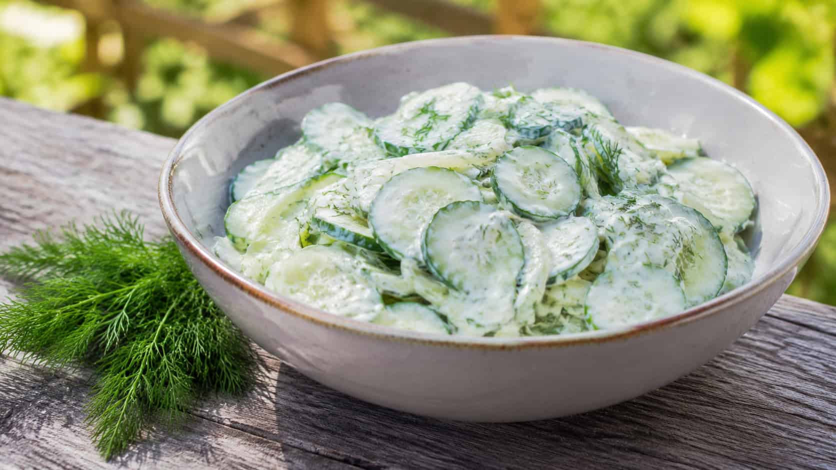 a creamy cucumber dill salad with thin cucumber slices and fresh dill in a serving bowl, shown in a wide horizontal shot on a picnic table with a sprig of dill nearby, natural light, shallow depth of field, landscape orientation