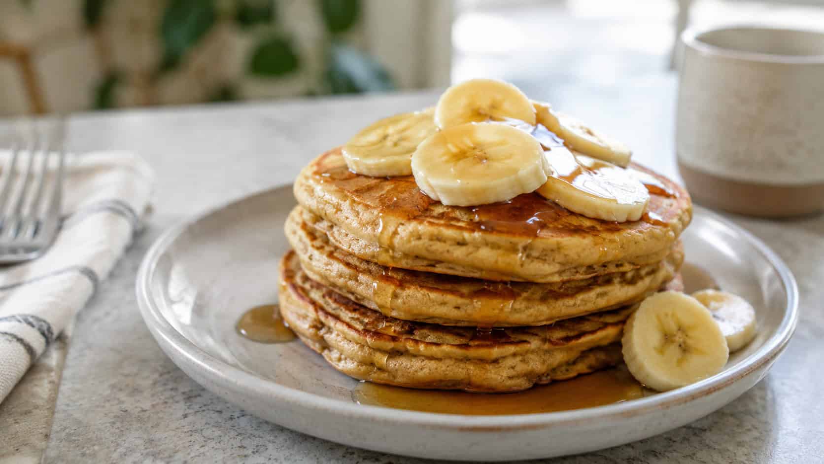 a short stack of golden banana pancakes topped with fresh banana slices and a drizzle of syrup, captured in a wide horizontal shot on a ceramic plate over a light table, natural light, shallow depth of field, landscape orientation
